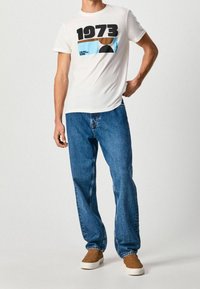 Mand iført hvid grafisk "1973" t-shirt, blå jeans og brune slip-on sko, der står mod en lys ensfarvet baggrund.