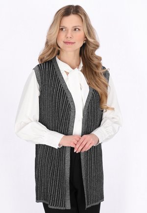 Femeie cu păr blond lung, purtând o bluză albă cu cravată și un cardigan fără mâneci în dungi negre și gri.