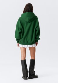 Felpa grande verde con cappuccio regolabile, tessuto texture, polsini a costine, indossata sopra una gonna bianca plissettata, abbinata a stivali neri fino al ginocchio.