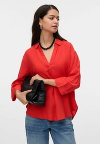 Blouse rouge, ample, avec un col en V et des manches trois-quarts, assortie à un jean bleu et une pochette noire. Le tissu semble léger.