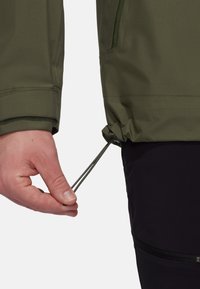 Oliwkowa kurtka o teksturowanym wykończeniu, regulowana na dole za pomocą linki ściągającej trzymanej w dłoni; częściowo widoczne czarne spodnie.