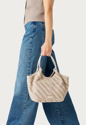 Mujer con vaqueros azules y top beige sosteniendo un bolso texturizado beige por sus dos asas a su lado.