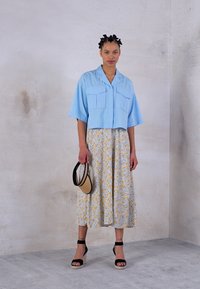 Camisa corta azul claro con dos bolsillos frontales, combinada con una falda larga floral en amarillo y azul, y sandalias de cuña negras con tiras.