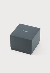 Boîte de montre Timex, forme carrée, en carton bleu marine foncé avec une finition mate. Présente un logo blanc en relief sur le dessus.