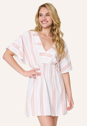 Blonde vrouw die glimlacht, gekleed in een korte, losse witte jurk met verticale lichtroze strepen en brede mouwen, hand op de heup.