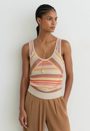 Femme aux cheveux tressés portant un haut en maille sans manches, rayé beige, rose et orange, un pantalon plissé beige et un long collier en or.