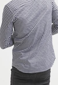 Camicia a quadretti vichy blu navy e bianca dal taglio aderente, con maniche lunghe e colletto ordinato, abbinata a jeans neri, vista da dietro.