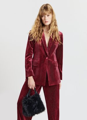 Red velvet blazer with a subtle dot pattern, paired with matching trousers. Model holds a small black fur handbag.