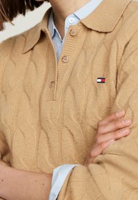 Pull en polo en tricot beige avec un col, doté d'une patte de boutonnage à trois boutons et d'une petite étiquette logo rouge, blanc et bleu.