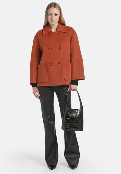 Manteau double boutonnage couleur orange brûlé avec de grandes poches, poignets de pull noir, porté sur un pantalon en cuir noir, tenant un sac à main en cuir noir gaufré crocodile.