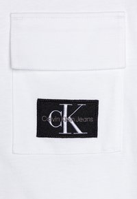 Biała koszulka z materiału z kieszonką naszywaną oraz czarną prostokątną metką z napisem "Calvin Klein Jeans" i białym logo "CK".
