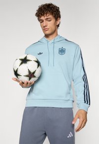 Light blue hoodie with a logo, dark blue side stripes, and a kangaroo pocket. Gray athletic pants and a black-and-white patterned soccer ball.