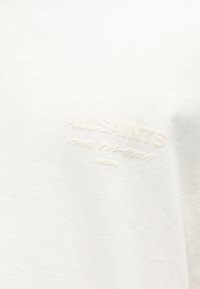 Κοντινό πλάνο λευκού υφάσματος με ανάγλυφα γράμματα που διαβάζουν «ALLSAINTS» και μικρότερους ιαπωνικούς χαρακτήρες από κάτω σε απλό φόντο.