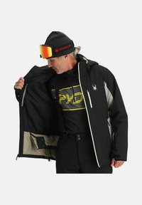 Schwarze isolierte Jacke mit hellgrauen Akzenten, innerer Reißverschlusstasche und reflektierendem Logo. Kombiniert mit einer schwarzen Mütze und Skibrille.