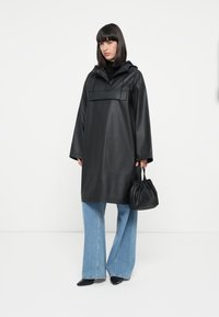 Black oversized raincoat with a hood, glossy finish, front pocket, paired with light blue wide-leg jeans and a small black handbag.