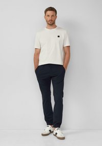 Witte katoenen t-shirt met een klein zwart vierkant logo, gecombineerd met donkerblauwe elastiche taillebroek en witte sneakers met bruine accenten.