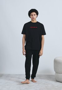 Čierne bavlnené tričko s vystupujúcim červeným logom „HUGO“, doplnené o čierne tepláky. Obe kúsky majú voľný strih a krátke rukávy.