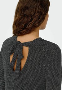 Zwart gestructureerd top met een patroonontwerp, open rug met een striksluiting en lange mouwen. De stof heeft een zachte, licht glanzende afwerking.