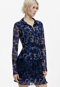 Blaues, floral bedrucktes Kleid mit langen Ärmeln, kragenähnlichem Ausschnitt, geschichteter, plissierter Rock und transparenter Textur. Verfügt über goldene Akzente.