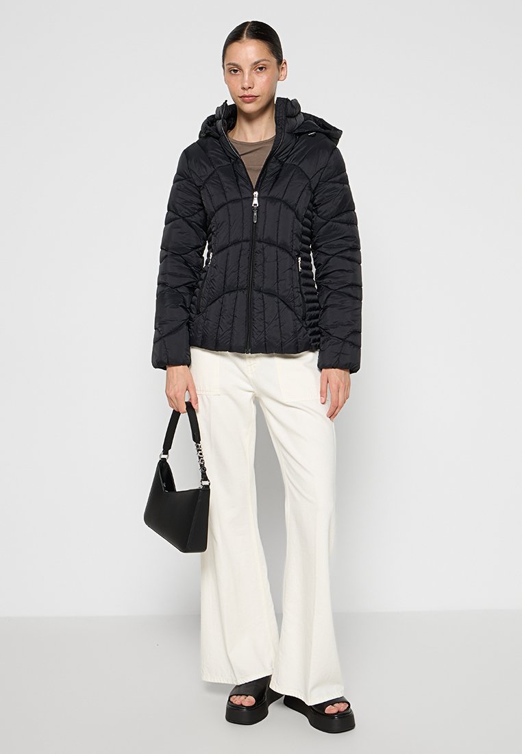 Chaqueta de plumas negra con capucha, con un diseño acolchado, cierre de cremallera y bolsillos laterales, combinada con pantalones acampanados blancos y un bolso negro.