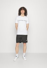 White cotton t-shirt with "Calvin Klein Jeans" logo, paired with black athletic shorts and white sneakers. Model stands against a plain backdrop.