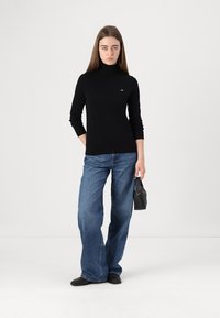 Pulover negru cu guler înalt, cu mâneci lungi, asortat cu blugi largi albaștri. Modelul ține în mână o poșetă mică neagră. Design simplu și curat.