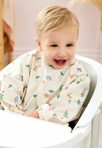 Hosszú ujjú bébielőke krém színben, színes dinoszaurusz és pálmafák mintáival, egy baba viseli, aki egy etetőszékben ül. Simára kidolgozott, vízálló anyag.