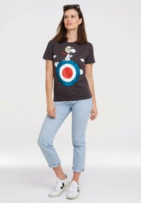 Gray t-shirt featuring Snoopy graphic on a blue and red target. Model wears light blue jeans and white sneakers with black accents.