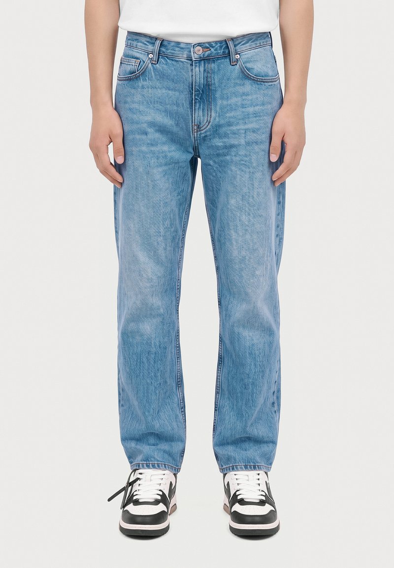 Personne portant un jean droit bleu clair et des baskets blanches et noires, debout avec les mains le long du corps.