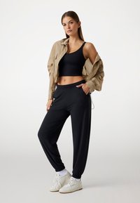 Crni crop top u kombinaciji s labavim crnim hlačama, laganom tan jaknom i bijelim tenisicama s teksturiranim detaljima i elastičnim rukavima.