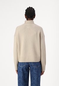 Beige, grob gestrickter Pullover mit hohem Kragen, gerippten Bündchen und Saum, kombiniert mit blauen Jeans im entspannten Schnitt, die mit floralen Mustern verziert sind.