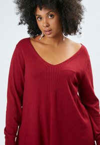 Burgunderfarbener Pullover mit langen Ärmeln und V-Ausschnitt, der über strukturierte Details auf der Brust verfügt und aus weichem Strickmaterial gefertigt ist.