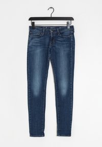 Jeans skinny de denim azul con una textura suave, diseño estándar de cinco bolsillos y un leve desgastado en los muslos. Cuelgan de un gancho negro.