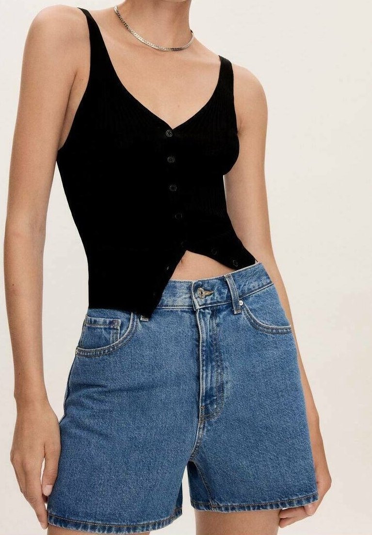 Femme portant un débardeur noir boutonné et un short en jean bleu taille haute, avec un collier chaîne argenté.