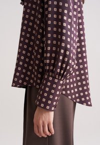 Langärmlige Bluse aus dunkelviolettem Stoff mit einem Rautenmuster aus kleinen beigen Quadraten. Verfügt über geraffte Manschetten und einen leicht asymmetrischen Saum.