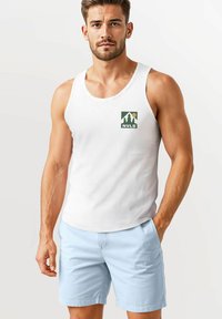 Weißes Tanktop mit einem kleinen grünen Logo, das Bäume und eine Sonne zeigt; kombiniert mit hellblauen Shorts aus glattem Material und seitlichen Taschen.