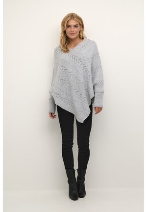 Hellgrauer gestrickter Poncho mit Zickzackmuster und asymmetrischem Saum, kombiniert mit schwarzen, eng anliegenden Hosen und Stiefeletten. Weiche Textur.