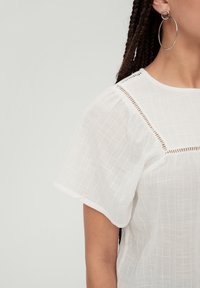 Hvid bluse med et tekstureret, gittermønster. Har korte, flagrende ærmer og diskrete åbne syninger over skuldrene for detaljer.