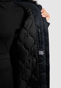 Schwarze gesteppte Steppjacke mit glatter Außenseite, Innentasche, Kapuze mit Kunstfellbesatz und Logopatch auf dem Innenfutter.