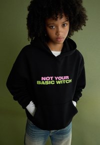 Czarna bluza z kapturem z przednią kieszenią, z napisem "NOT YOUR BASIC WITCH" w różowym i zielonym kolorze. Model nosi jasnoniebieskie dżinsy.