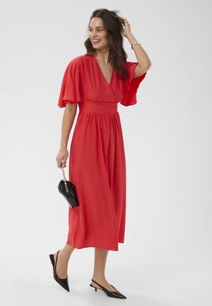Femme en robe midi rouge fluide à manches volantées, portant un petit sac à main noir et des escarpins noirs à talons bas, souriante et se touchant les cheveux.