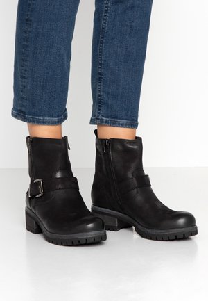 Bottines en cuir noir avec fermetures éclair latérales, brides bouclées, talons épais, portées avec un jean bleu retroussé sur un sol blanc.