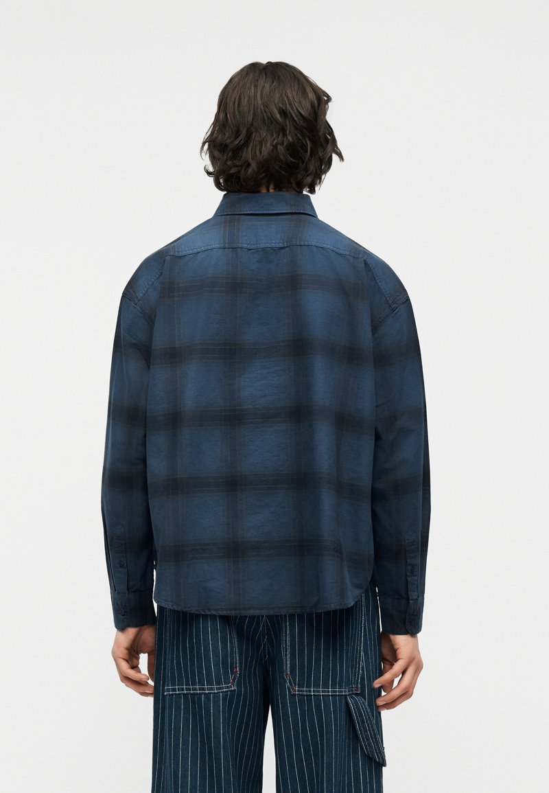 Homme aux cheveux mi-longs vu de dos, portant une chemise à manches longues à carreaux bleus et un pantalon bleu foncé à fines rayures avec une poche latérale.