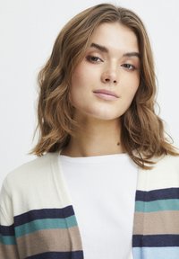 Adīta jaka ar horizontālām svītrām tumši zilā, teal, bēšā un krēmotā krāsā; atvērta priekšpuse; valkāta virs baltā T-krekla. Mīksta tekstūra, brīvs piegriezums.