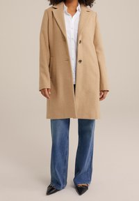 Beige kappa i ullblandning med ett snaggat krage, två knappar och sidofickor, kombinerad med en vit skjorta och blå utsvängda jeans. Svarta spetsiga skor.