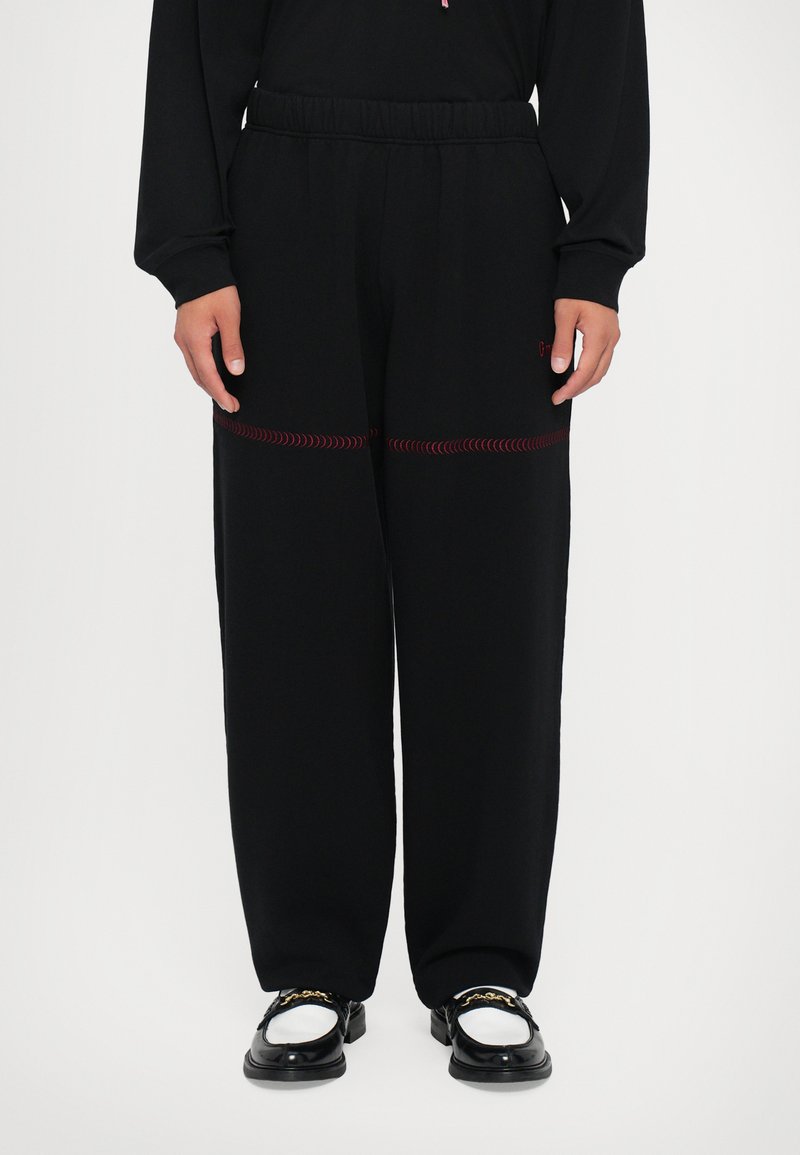 Pantalones de chándal negros con un ajuste relajado, con costuras en zigzag rojas a lo largo de las piernas y una cintura elástica. Combinados con zapatos negros.