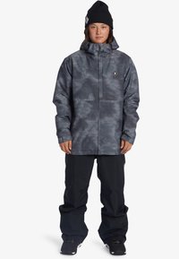 Šedá maskovací bunda se zipovým zapínáním, obsahující kapuci a nášivku s logem, v kombinaci s volnými černými kalhotami a černými botami.