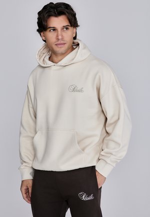 Cremefarbener Hoodie mit Fronttasche und besticktem Logo, kombiniert mit braunen Hosen mit weiß gesticktem Logo. Weiche Textur, lockere Passform.