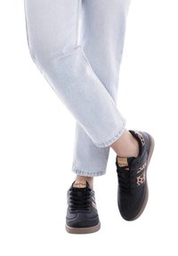 Baskets noires avec des accents en imprimé léopard et des semelles en caoutchouc marron, associées à un jean cropped bleu clair. Lacets et matériau texturé visibles.