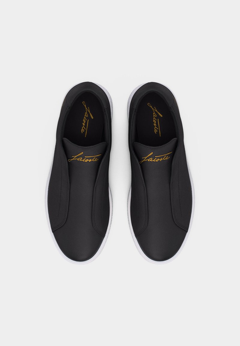 Coppia di sneakers nere senza lacci con suole bianche e logo dorato "Lacoste" sulla linguetta e sulle solette, mostrata vista dall'alto su sfondo bianco.